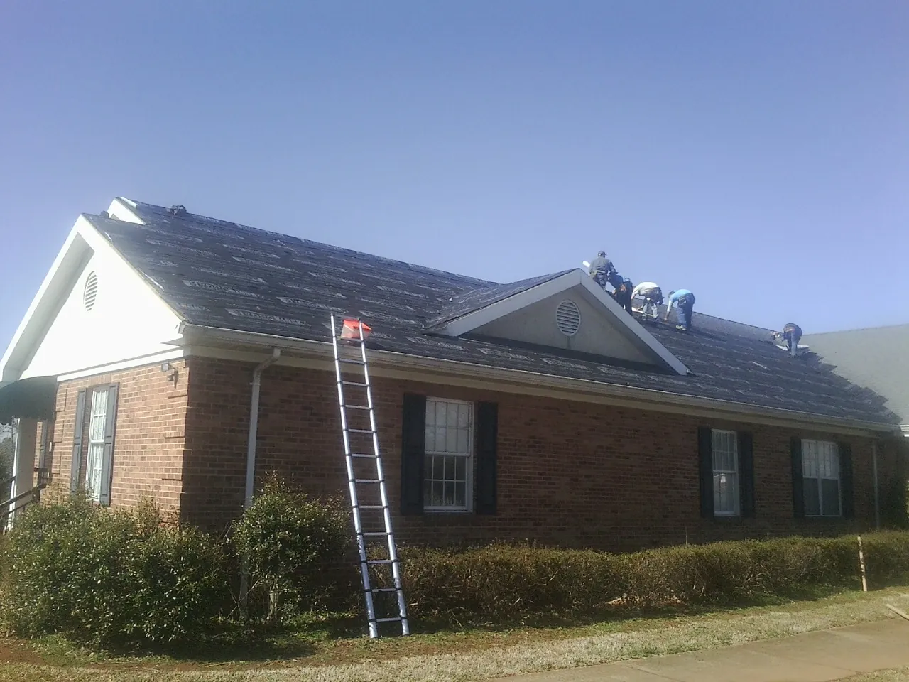 Crew tearing shingles off a brick ranch roof with a ladder set — Emergency Roof Repair work by our crew in Dana