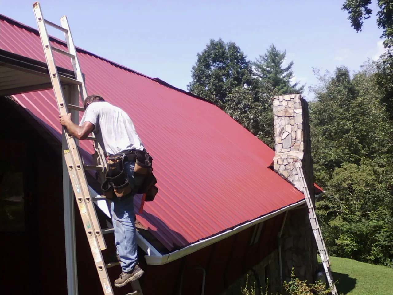 Roofer on a ladder beside a red metal roof and stone chimney — Soffit & Fascia Repair by Roofing Brothers in Dana